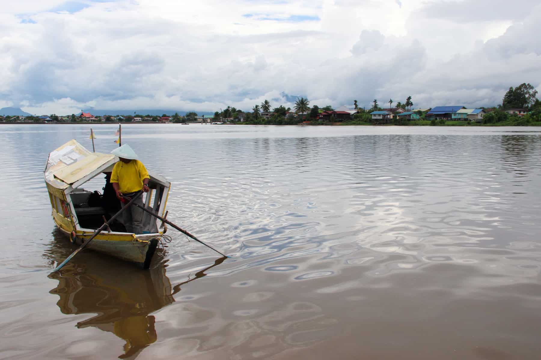 A man in a yellow t-shirt rows a white roofed boat with wooden oars on the chocolate brown waters of the Sarawak River in Kuching