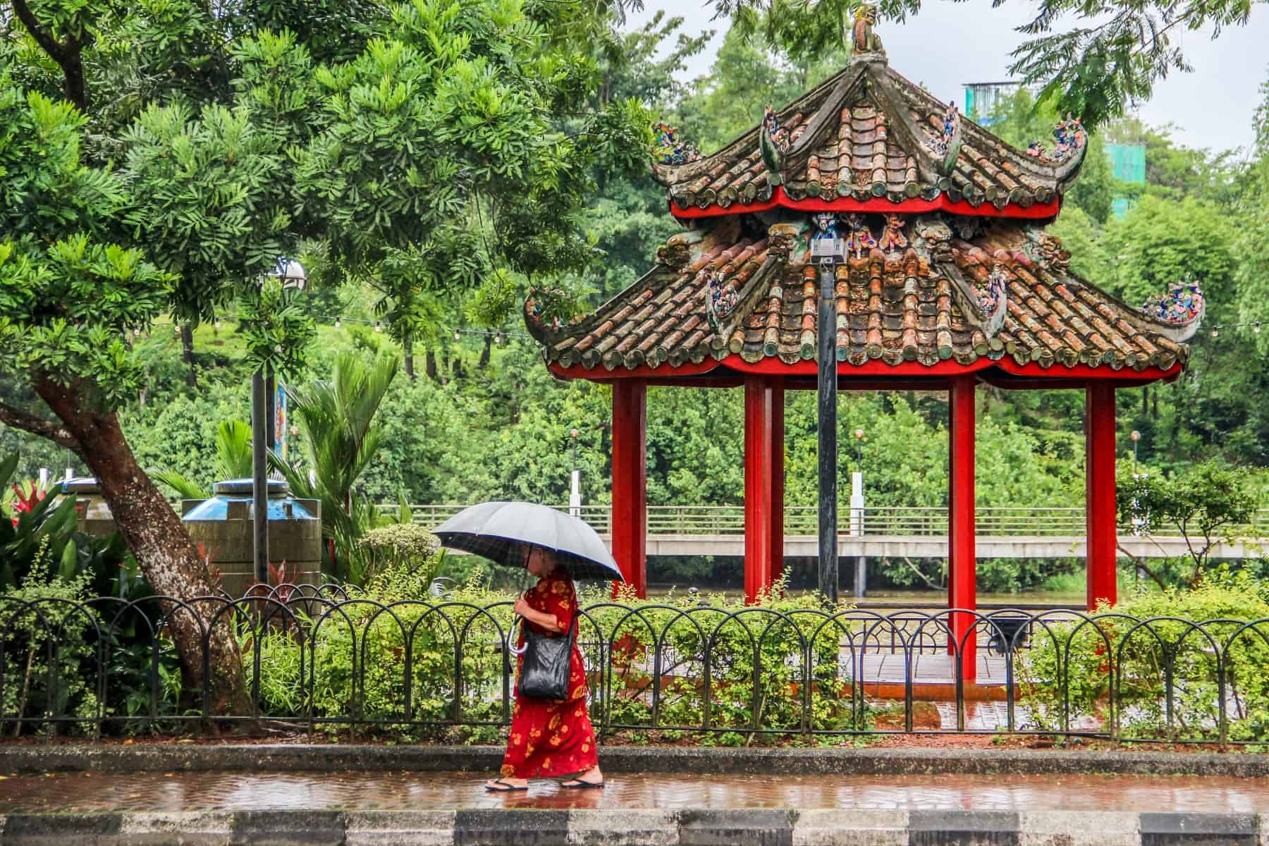 A lady in red, holding a sun umbrella walked past a striking red Chinese Pagoda set within bold green woodland along a riverbank in Kuching