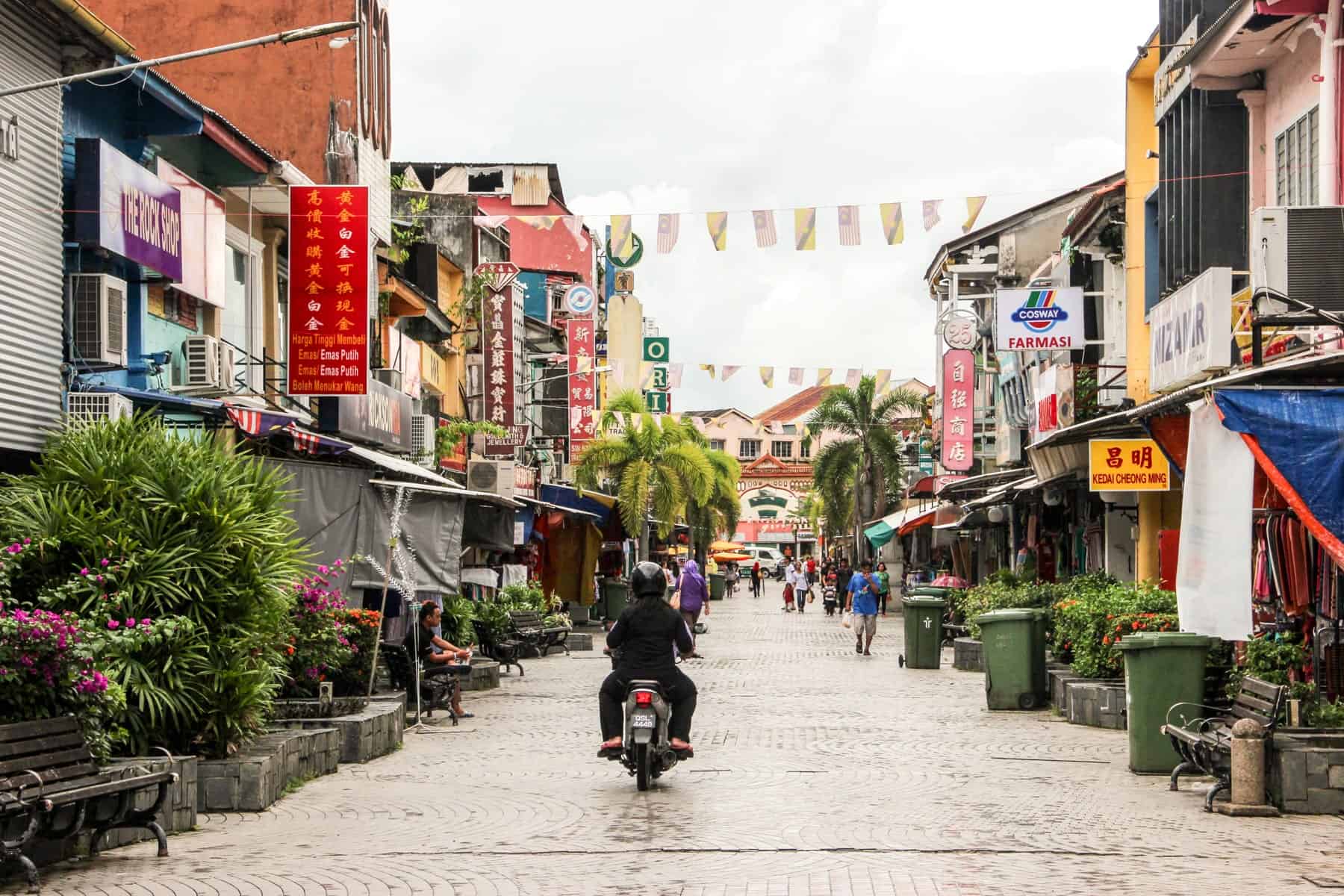 A man rides a motorcycle down a paved street in Kuching Sarawak, lined with local stores and decorated overhead with colourful bunting