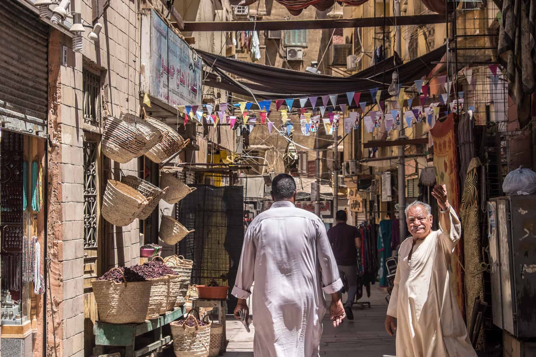 An older man in a white tunic waves in a souk in Aswan, Egypt. Behind him in a souk alleyway filled with shops whose doorways are lined with products