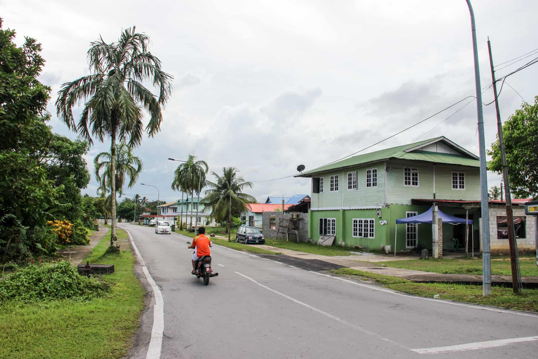 A man in an organe t-shirt rides a motor bike on a paved road past a mint green wooden house in a local neighbourhood in Kuching