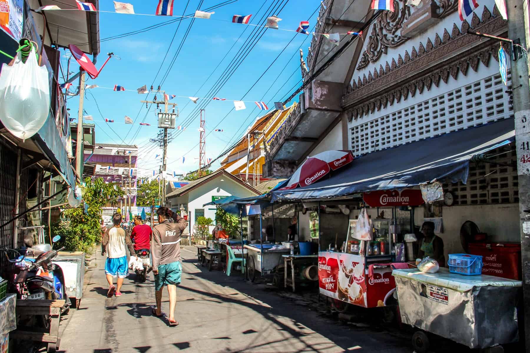 Three young men walk through a market stall lined street in Outer Bangkok in the midday sun