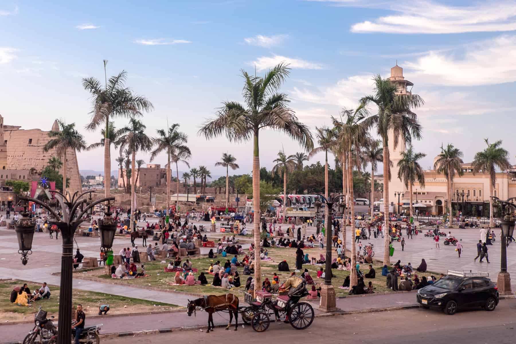 A public square in front of Luxor Temple in Egypt filled with palm trees, horse and. carts, crowds of people