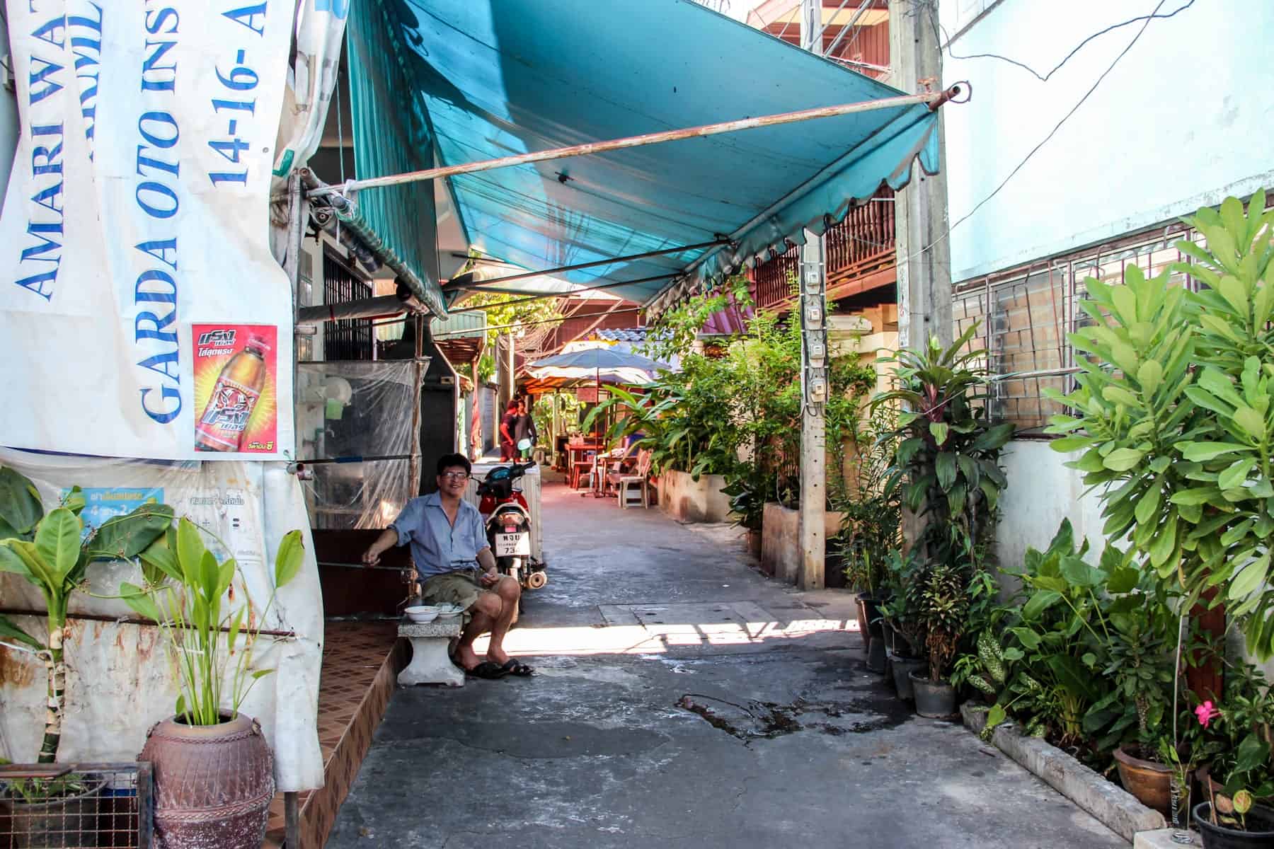 A Thai man sitting outside his market stall in Outer Bangkok under the shade of a blue canope