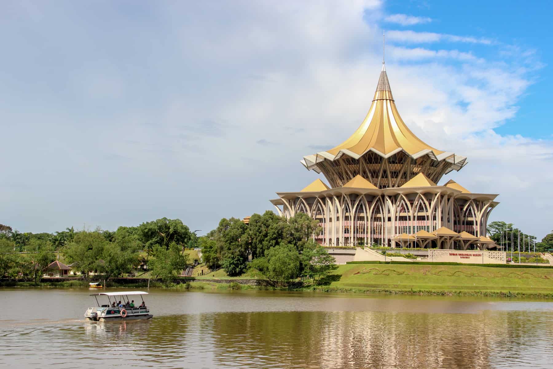 A boat glides past the golden two-tiered cake-like structure with a pointed hat style roof of the New Sarawak State Legislative Assembly Building in Kuching, Borneo