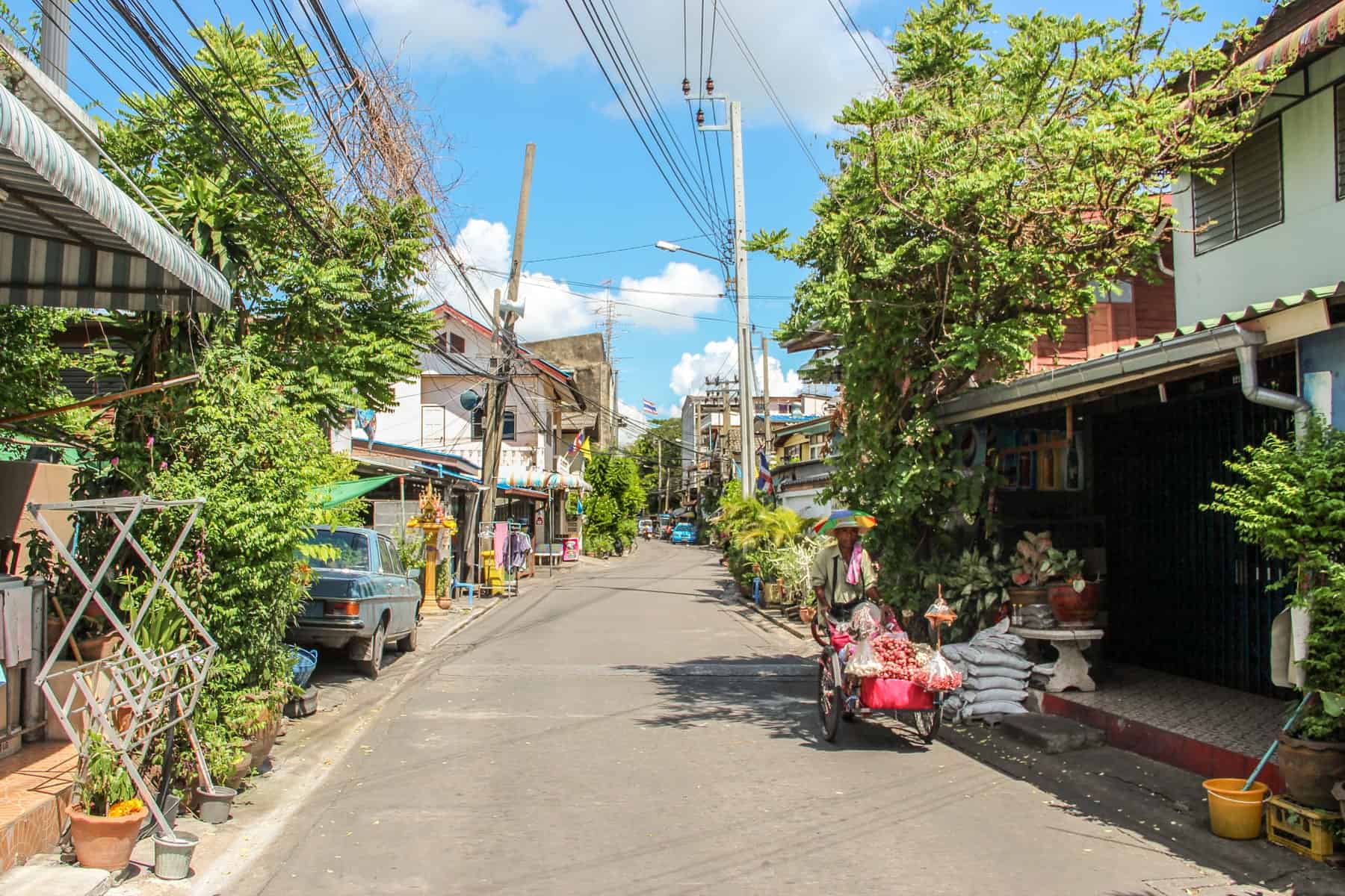 A man in a multicoloured sun hat pushes a wheelbarrow of fruit in a leafy, tree lined neighbourhood lined on both sides with white houses, in Outer Bangkok