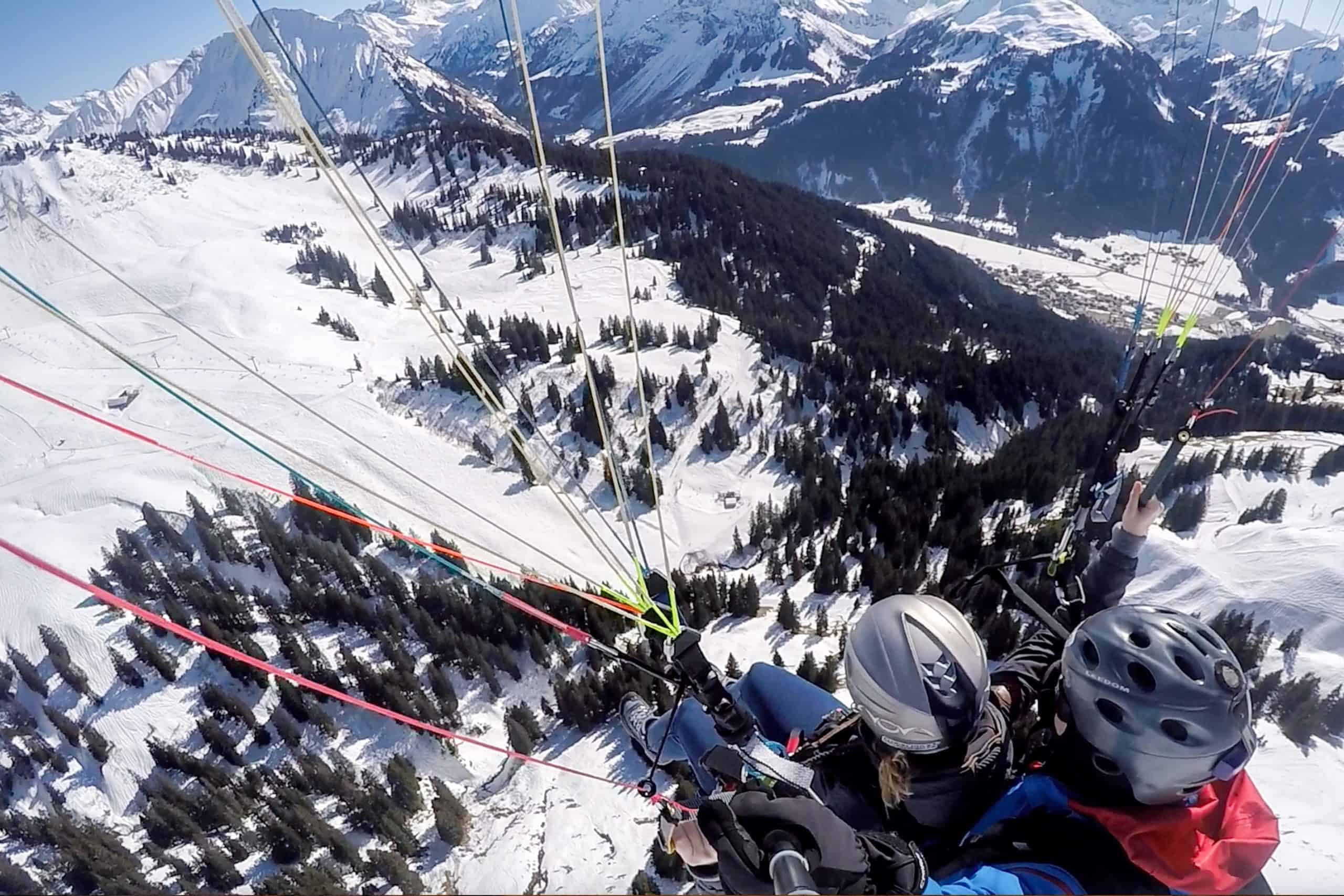 The two helmets and parachute strings of a man and woman in tandem paragliding soaring over a snowy, tree filled valley in Vorarlberg Austria