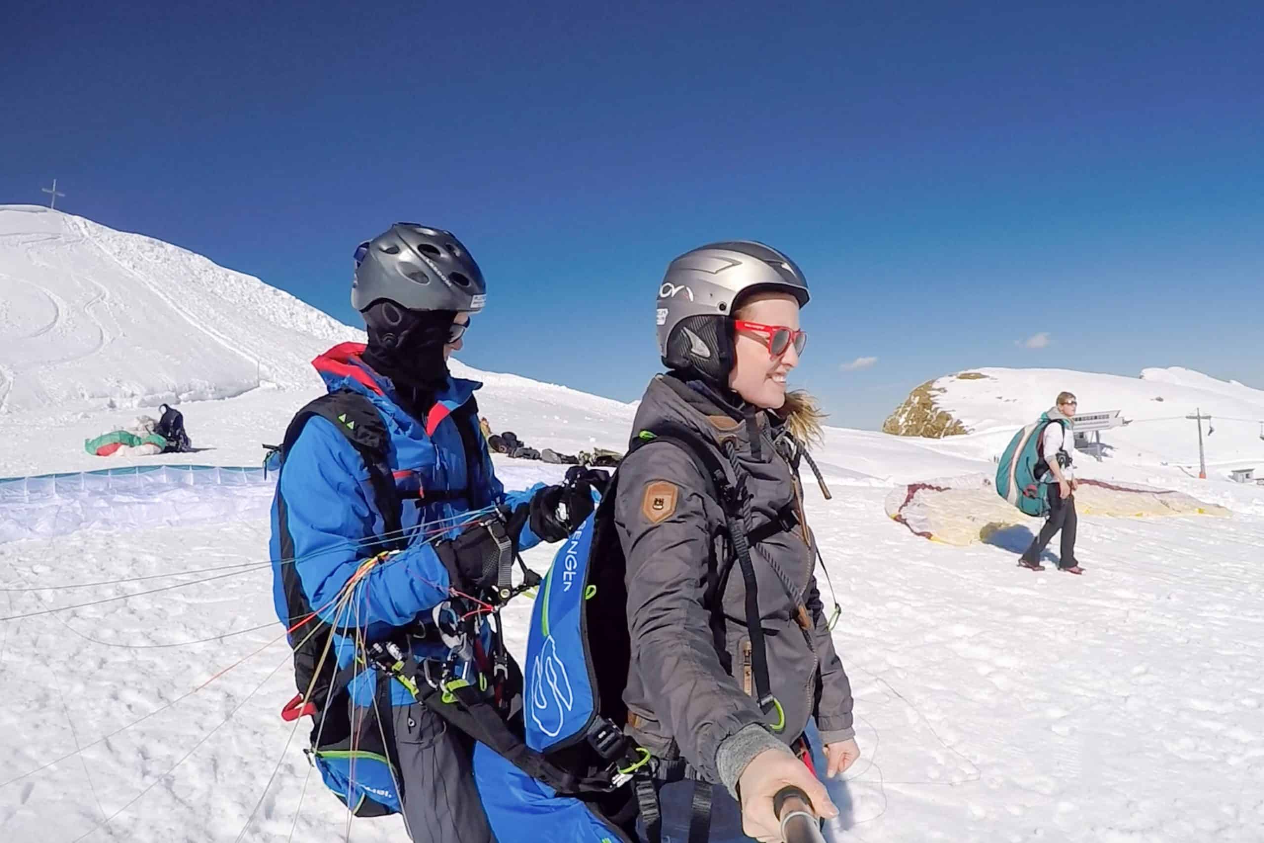 A male paragliding pilot setting up a woman's harness on a mountain in Austria