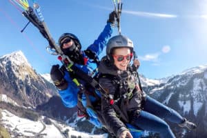 A paragliding pilot in bright blue twirls a woman passenger in the air over the Austrian Alps