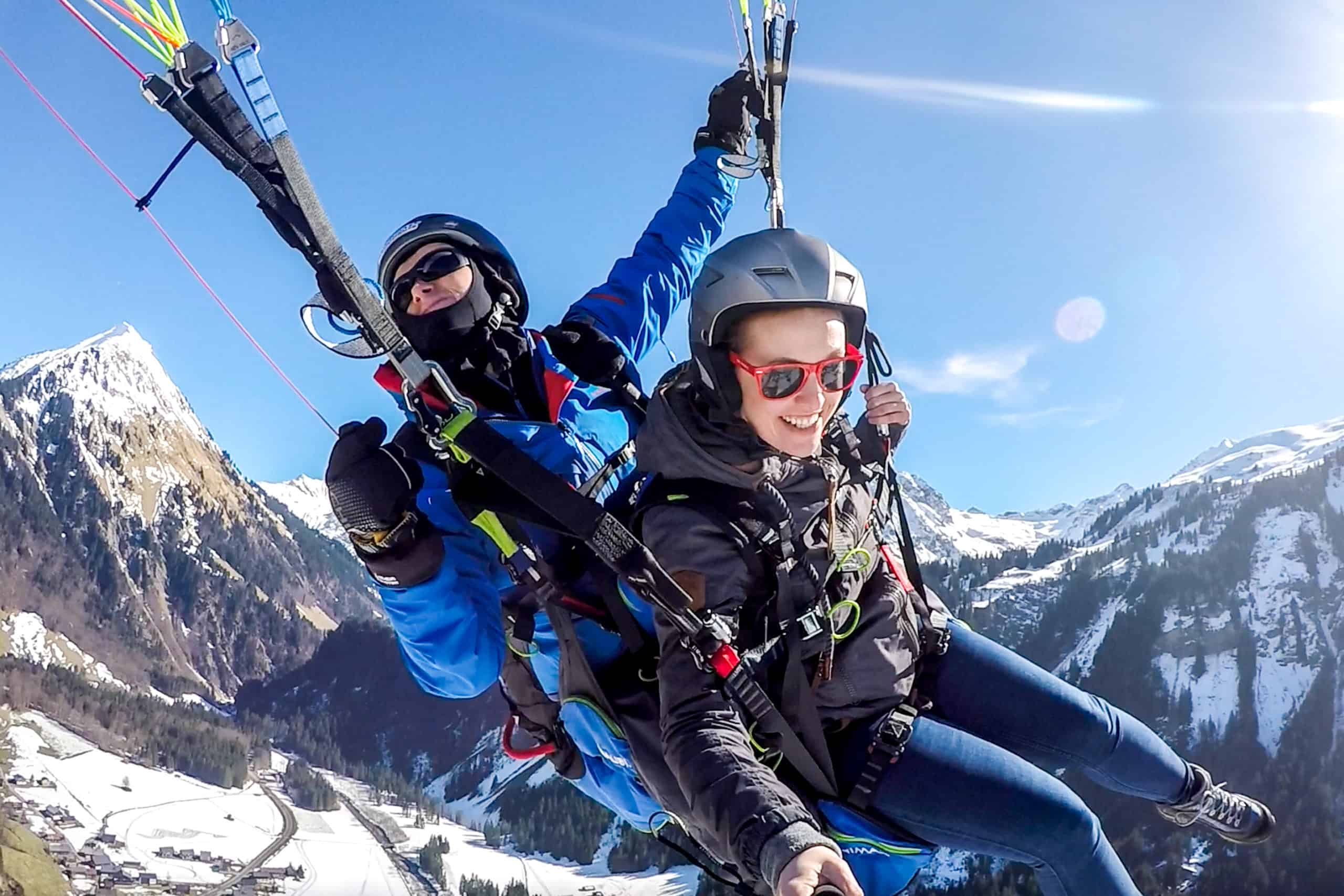 A paragliding pilot in bright blue twirls a woman passenger in the air over the Austrian Alps