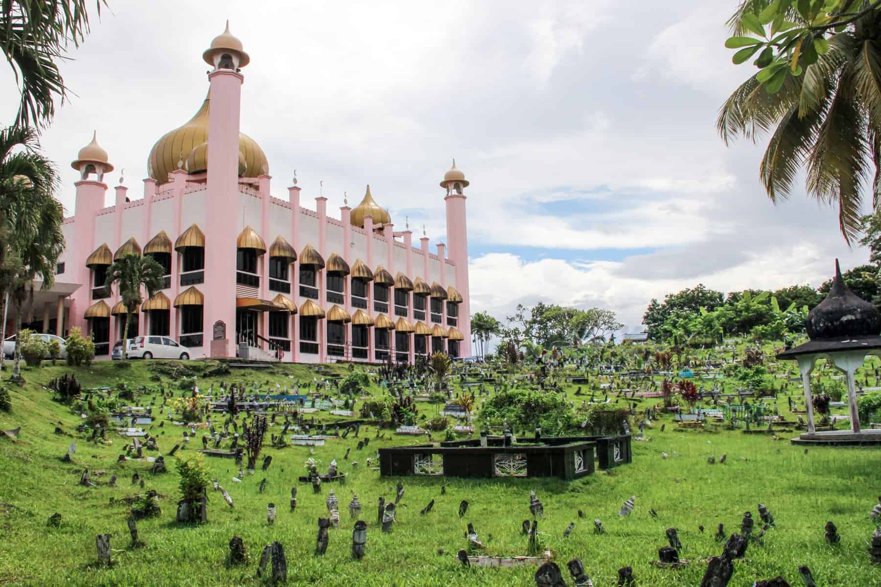The two-window tiered pink building and golden minarets of the Petra Jaya Old State Mosque Kuching, Sarawak. It stands within a large green area where small stone tomb stones poke out of the grass.
