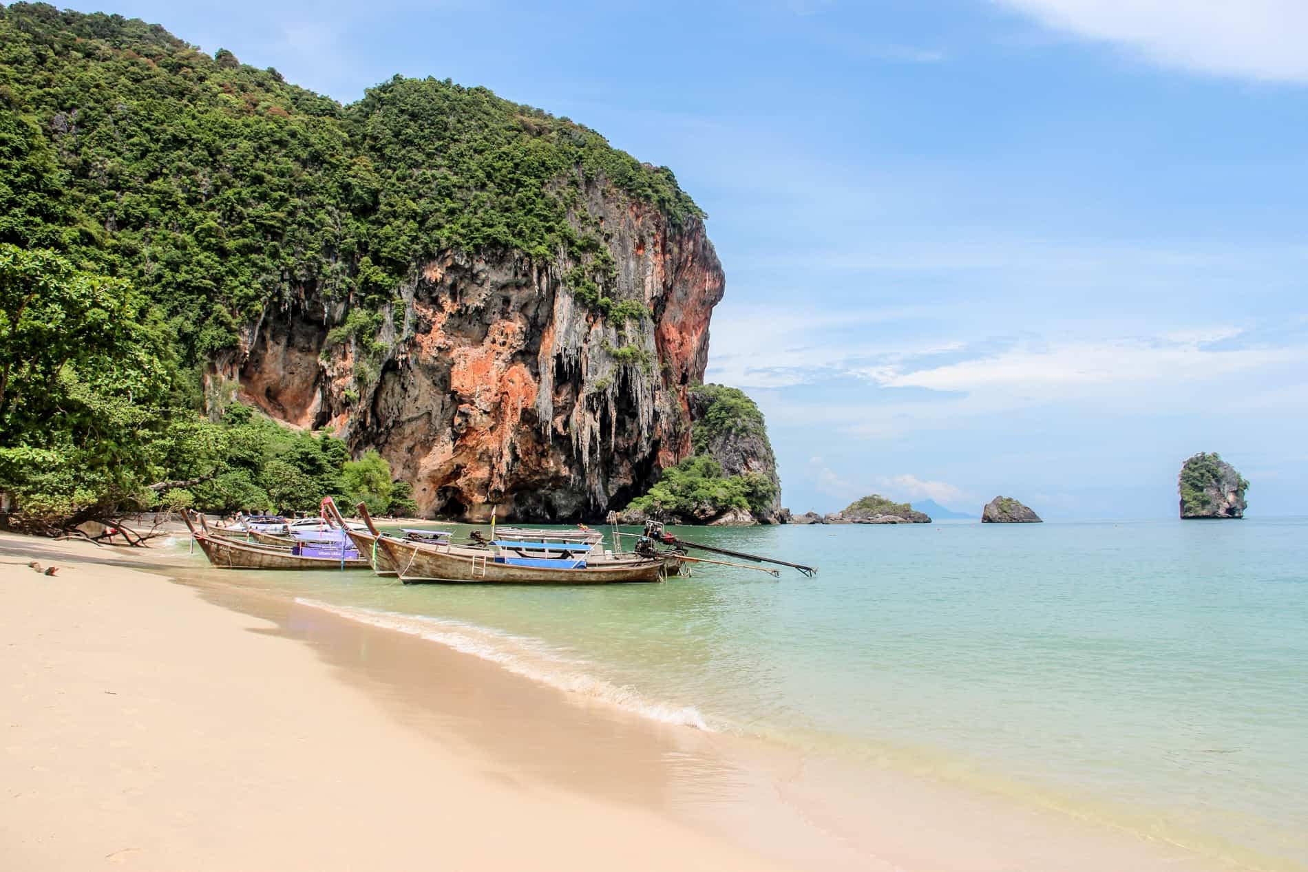 Wooden boats moored at yellow sand Railey Beach in Krabi, next to tree-covered limestone cliffs. 