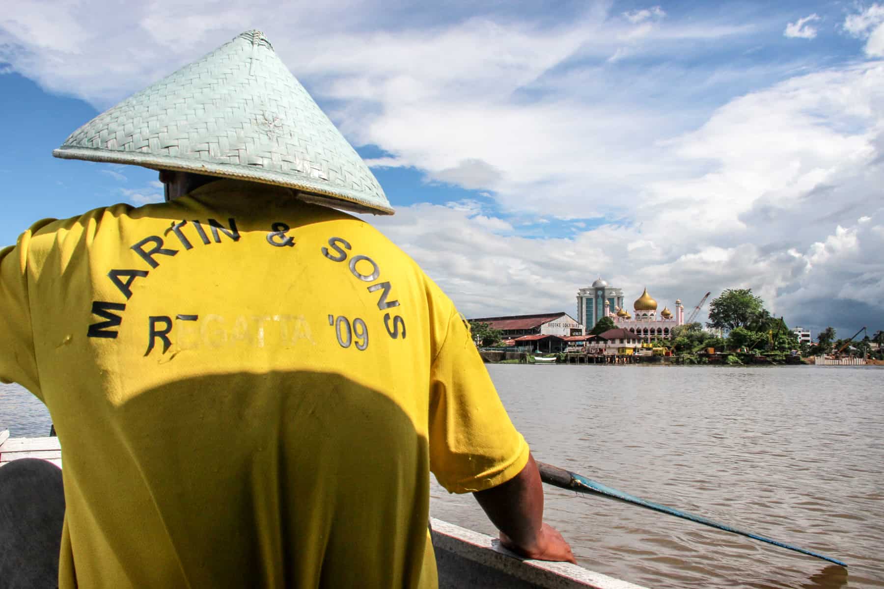 A man in a yellow t-shit and a triangular hat, uses a wooden oar to row across the River Sarawak in Kuching as a water taxi service