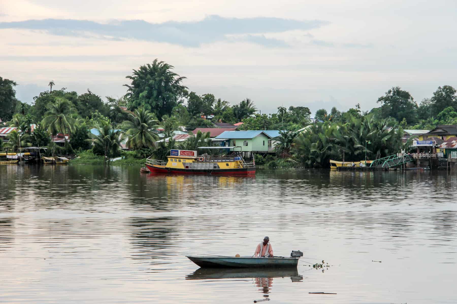 A man rows a small wooden boat on the Sarawak River in Kuching. In the background is the local neighbourhood marked by rows of colourful painted houses in bright colours, set within jungle green