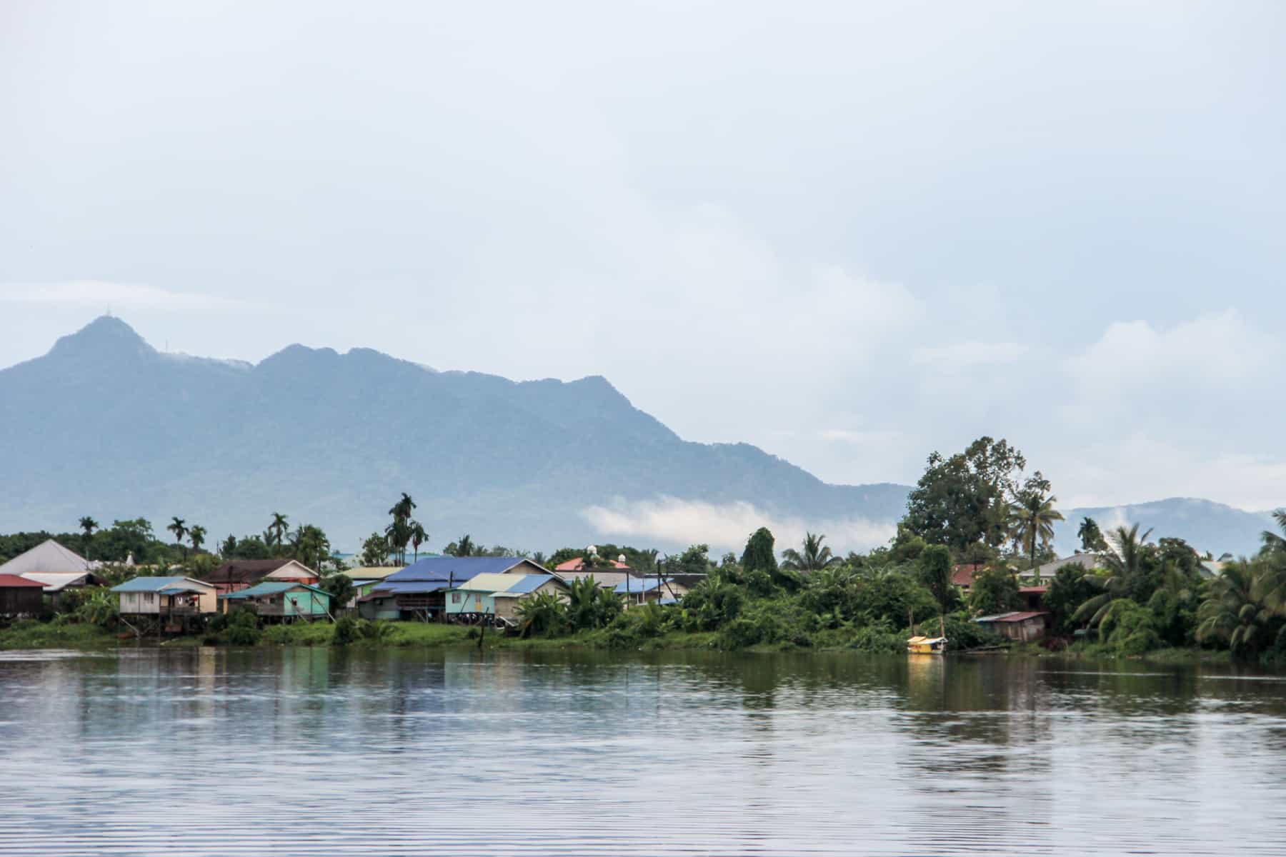 A row of multicoloured wooden houses line the Sarawak River in Kuching, to a background of mountains and lush greenery