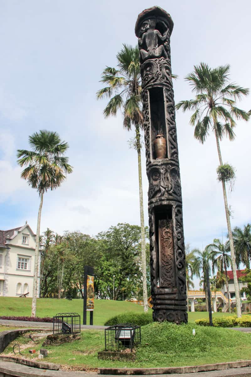 The weathered dark wooden totem pole stands on a green lawn amongst palm trees outside the white building of the Sarawak State Museum in Kuching