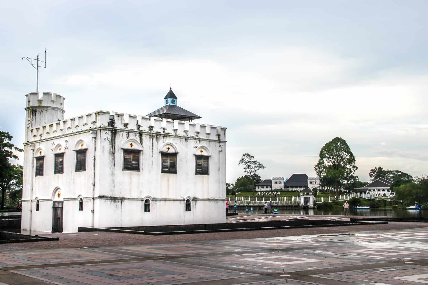 The white boxed structure of the Square Tower in Kuching resembles a mini fortress with turrets. It stands on the riverfront with The Asana old palace building in the background, covered in foliage