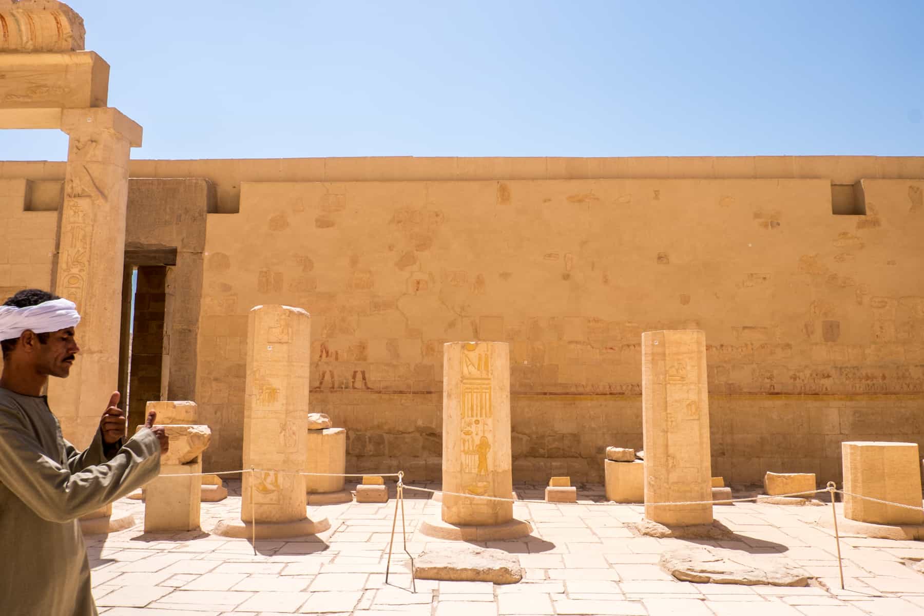 An Egyptian man in green tunic and white headband giving thumbs up to a tourist at a temple site in Luxor, Egypt