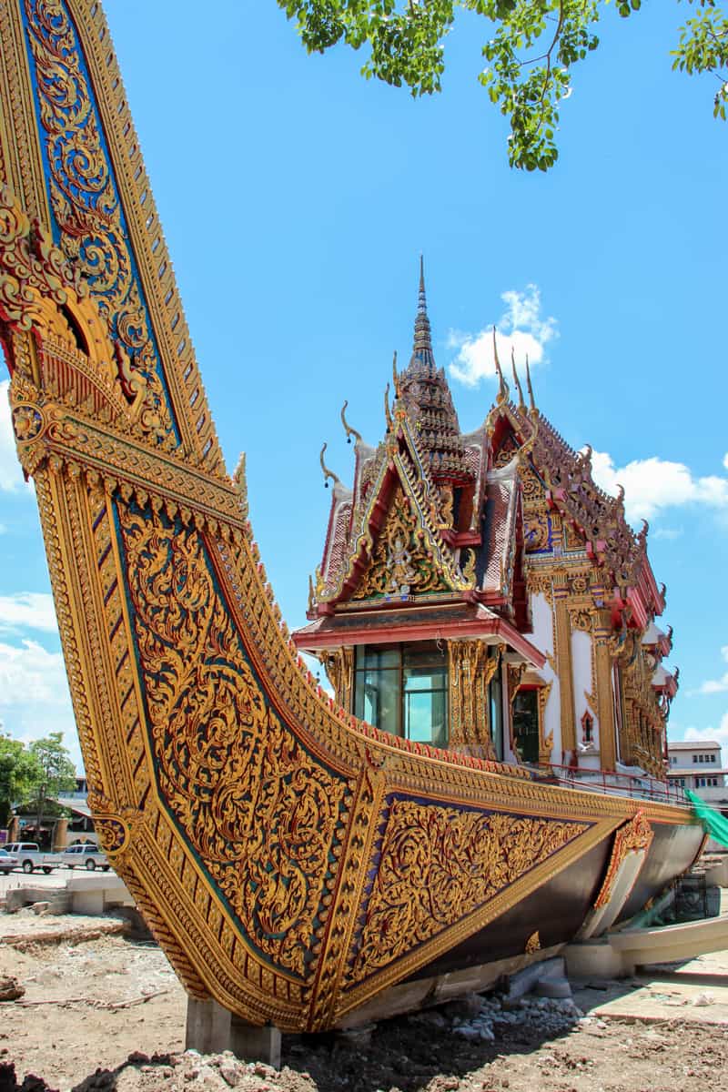 A triangular layered, red and gold decorated Thai temple structure sits in the middle of a giant golden boat structure in Outer Bangkok