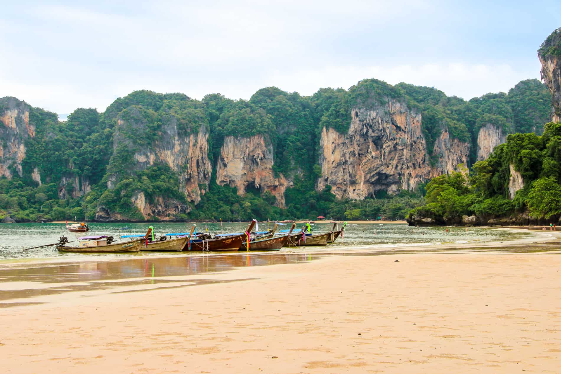 Wooden long boats docked on a limestone cliff-backed beach in Krabi, Thailand.