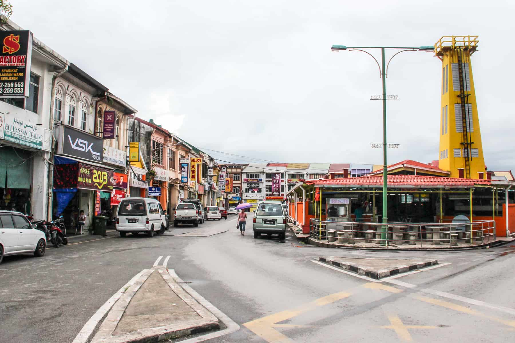 Next to a row of shops in colonial style architecture, an orange roofed building with a tall yellow tower sticking out of it is the site of The Old Fire Station in Kuching