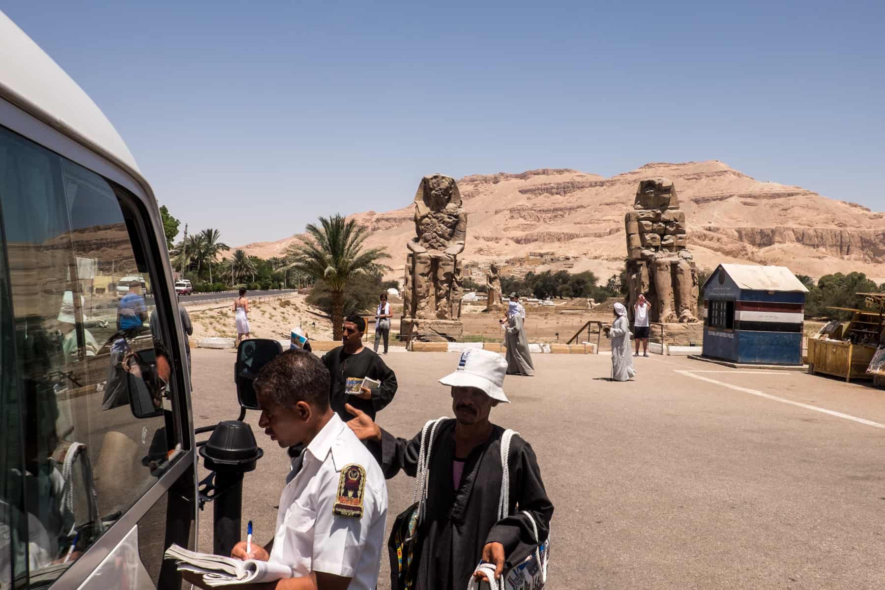 The Egyptian Tourism and Antiquities Police checking a white tourism van at an ancient statue site, with vendors behind selling handicrafts.