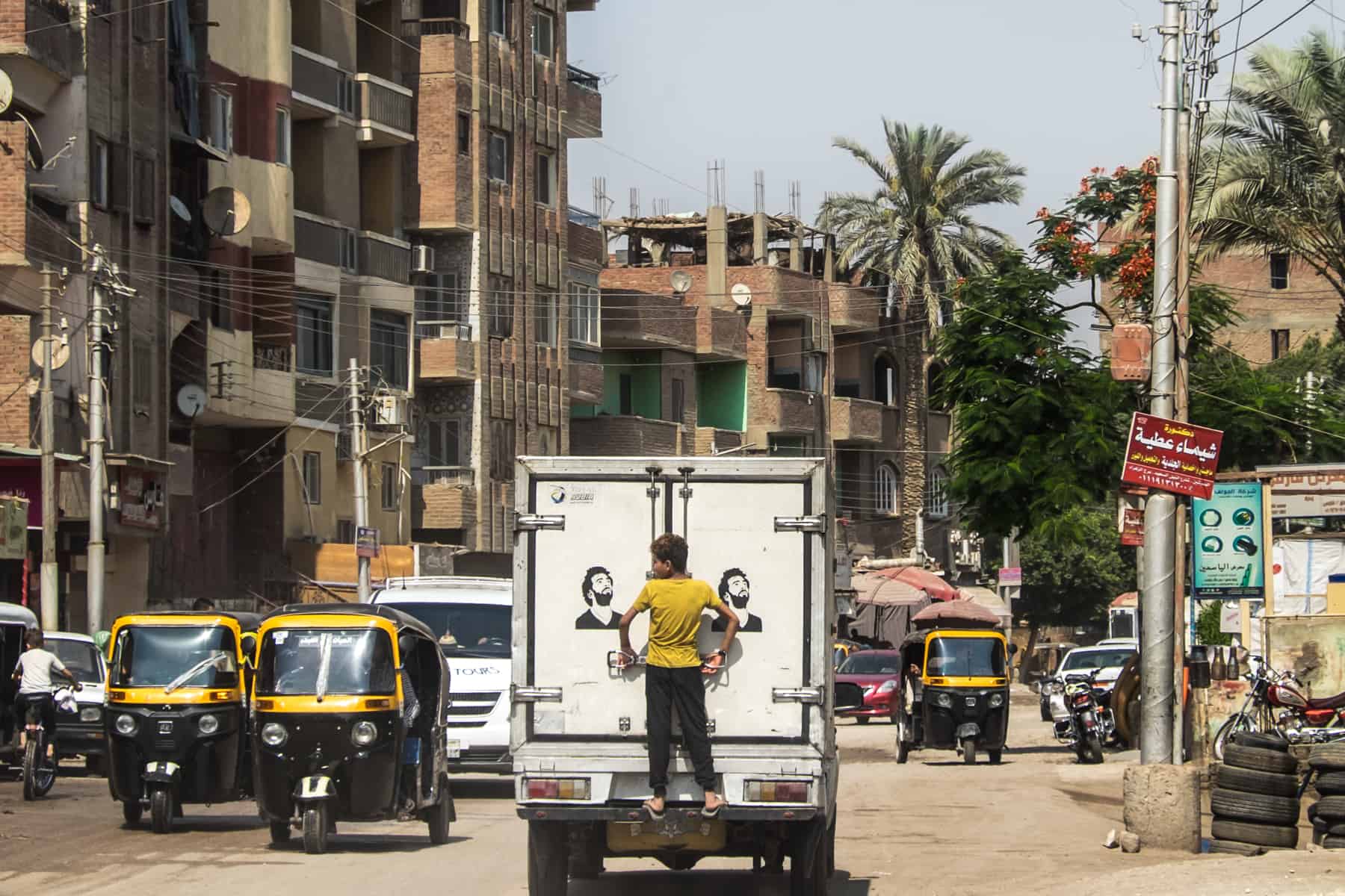 A young boy hangs off the back of a white van in Egypt, a free ride on a busy road. Black and yellow tuk tuks pass the van.