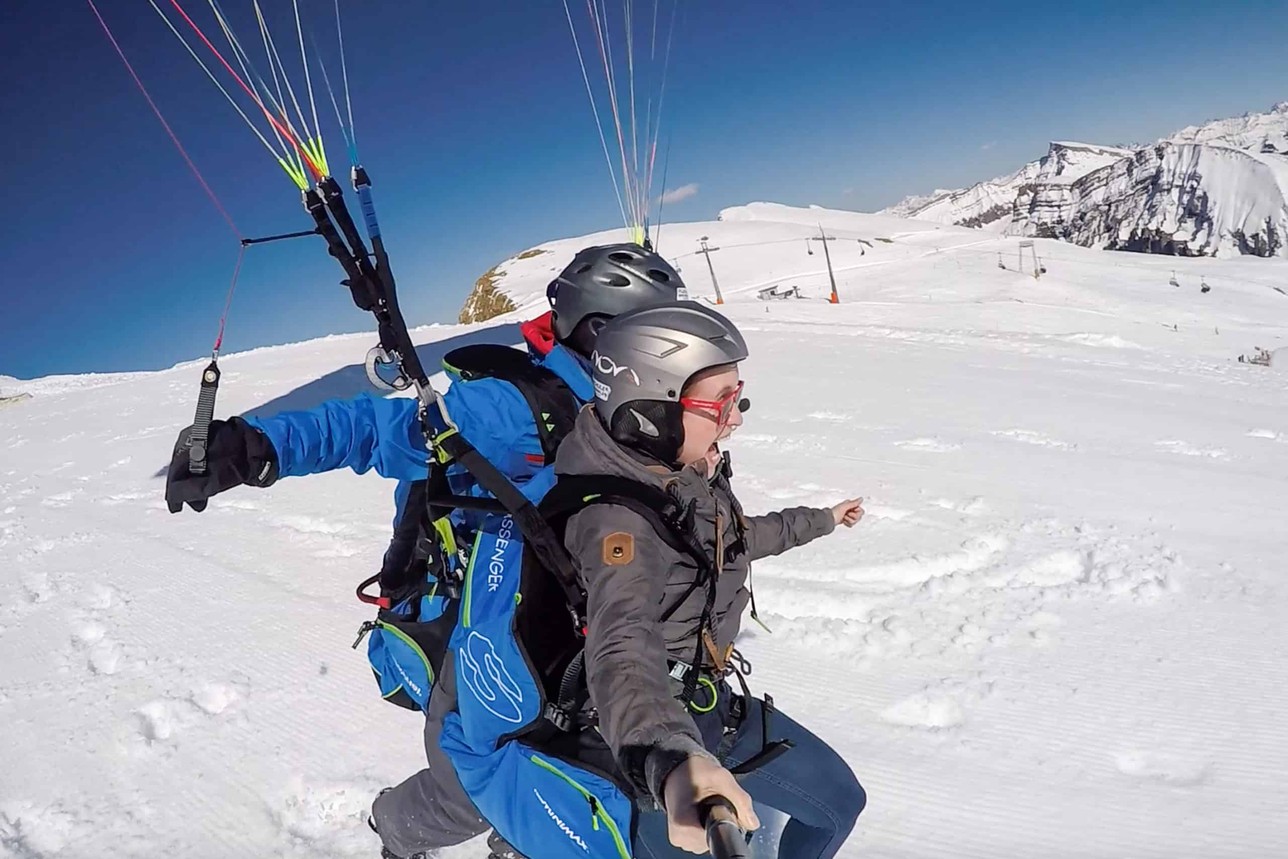 A woman with an excited facial expression tandem paraglides with a male pilot of a snowy mountain peak in Austria