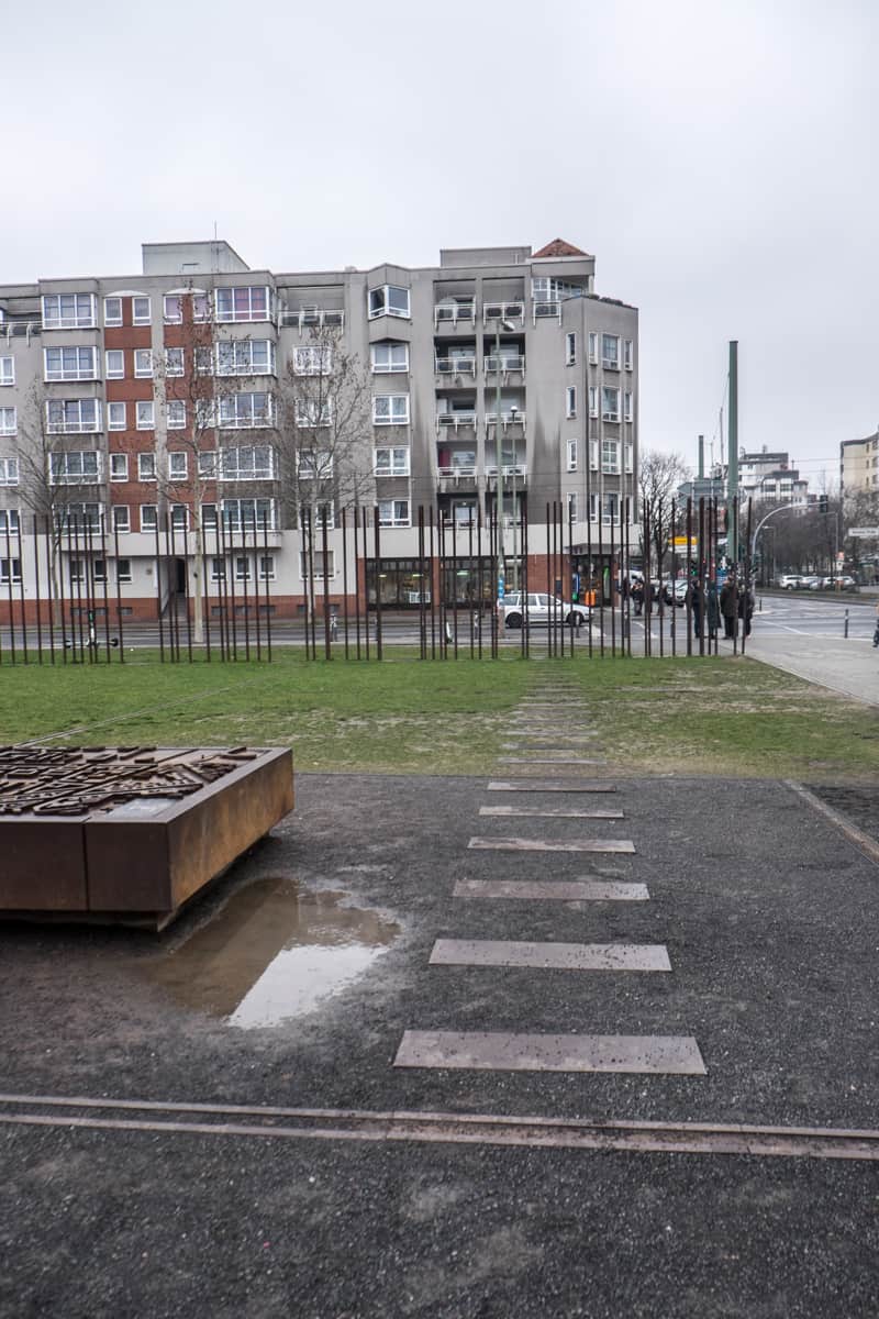 Rectangular panels on pavement and in the grass showing the route of an escape tunnel in Berlin, leading to the site of the Berlin wall, now marked by tall, bronze poles.