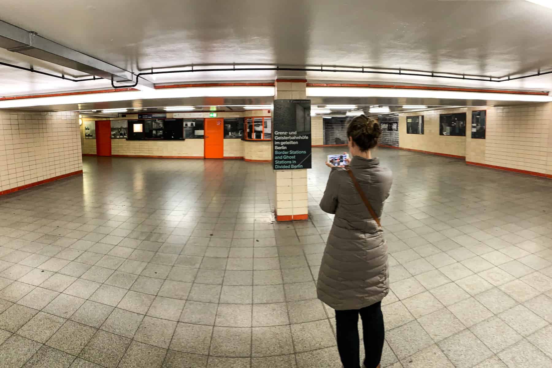 A woman in a brown coat stands in the middle of a beige tiled metro station in Berlin taking photos of her phone of the Border Stations exhibition images on the walls