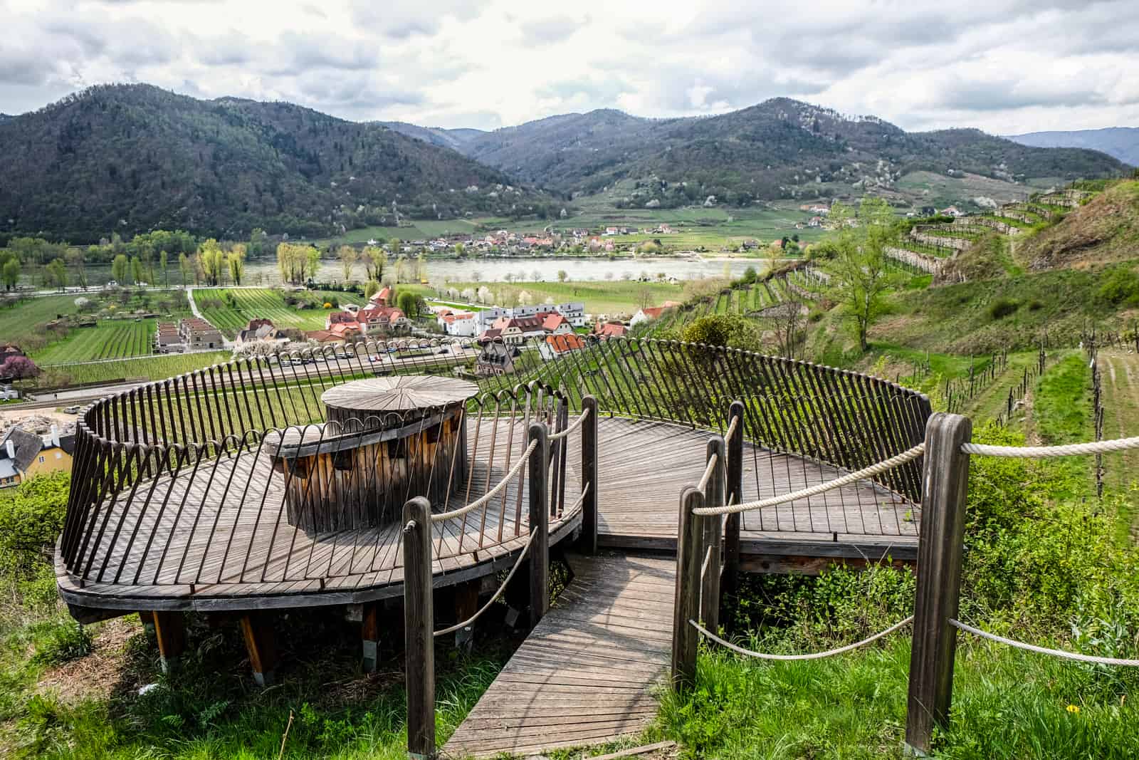 A snail shaped wooden platform with a chair sits on a green grassy hill overlooking the Danube River and the dots of red roofed village houses