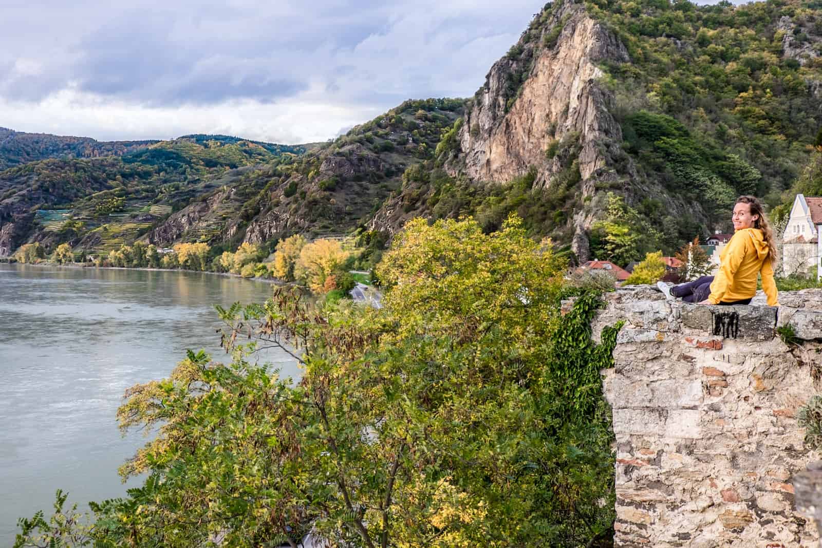 A woman wearing yellow sits on an old stone wall in the Wachau Valley next to the Danube River. She looks at the camera and smiles. Behind her is a large cliff face and a small white house.