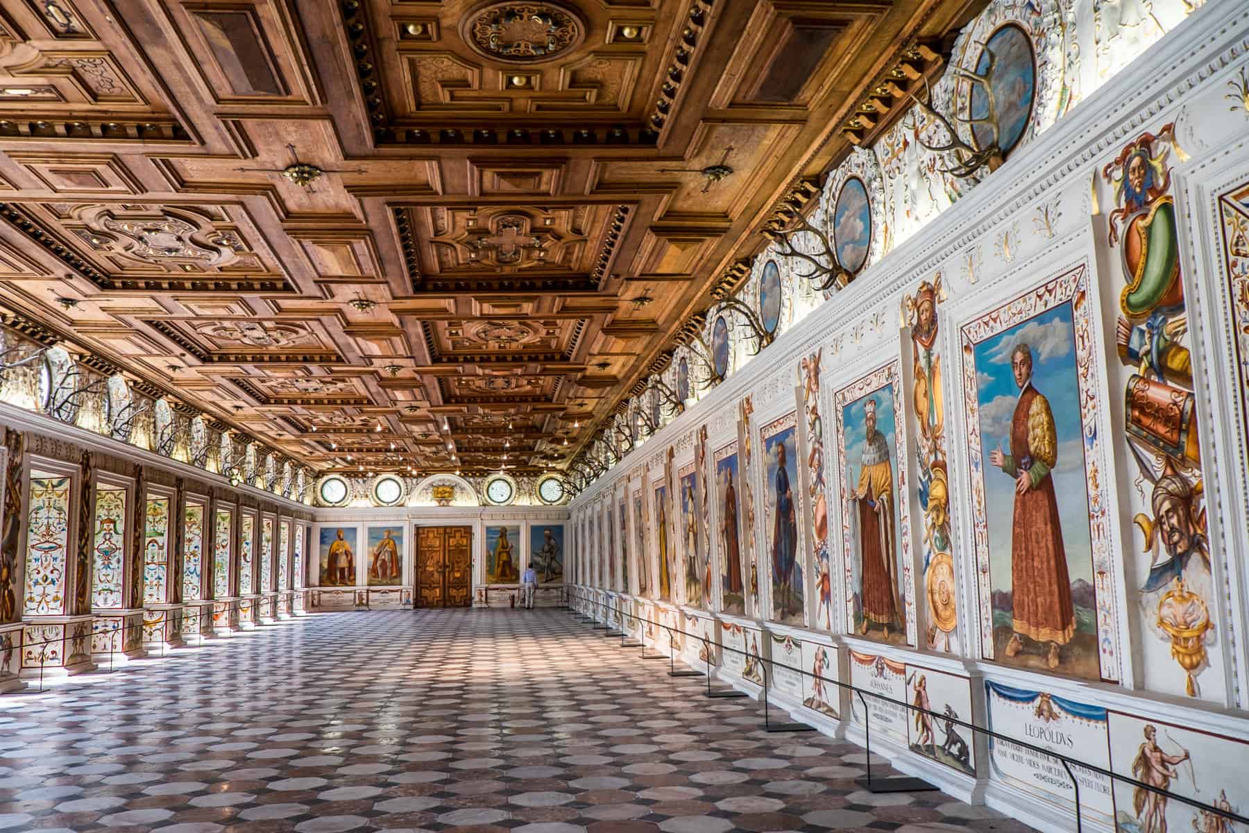 A wooden ceiling, tiled floored and old wall paintings of rulers line a grand hallway in Innbruck's Ambras Castle