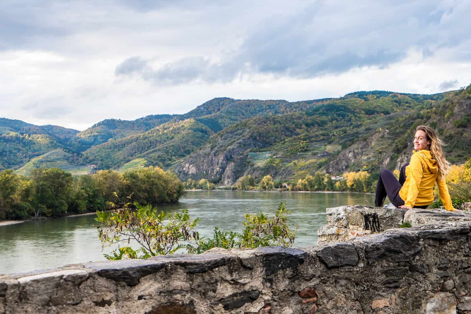 A woman is a yellow jacket sitting on the stone walls of the Wachau Valley Austria overlooking the Danube River