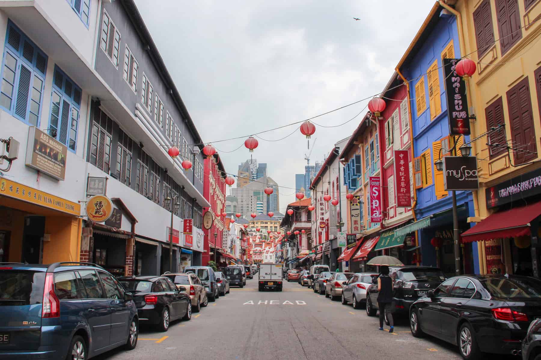 Red chinese lanterns line the middle of a long street in Singapore's Chinatown with colourful old buikdings on the right leading down to highrise buildings