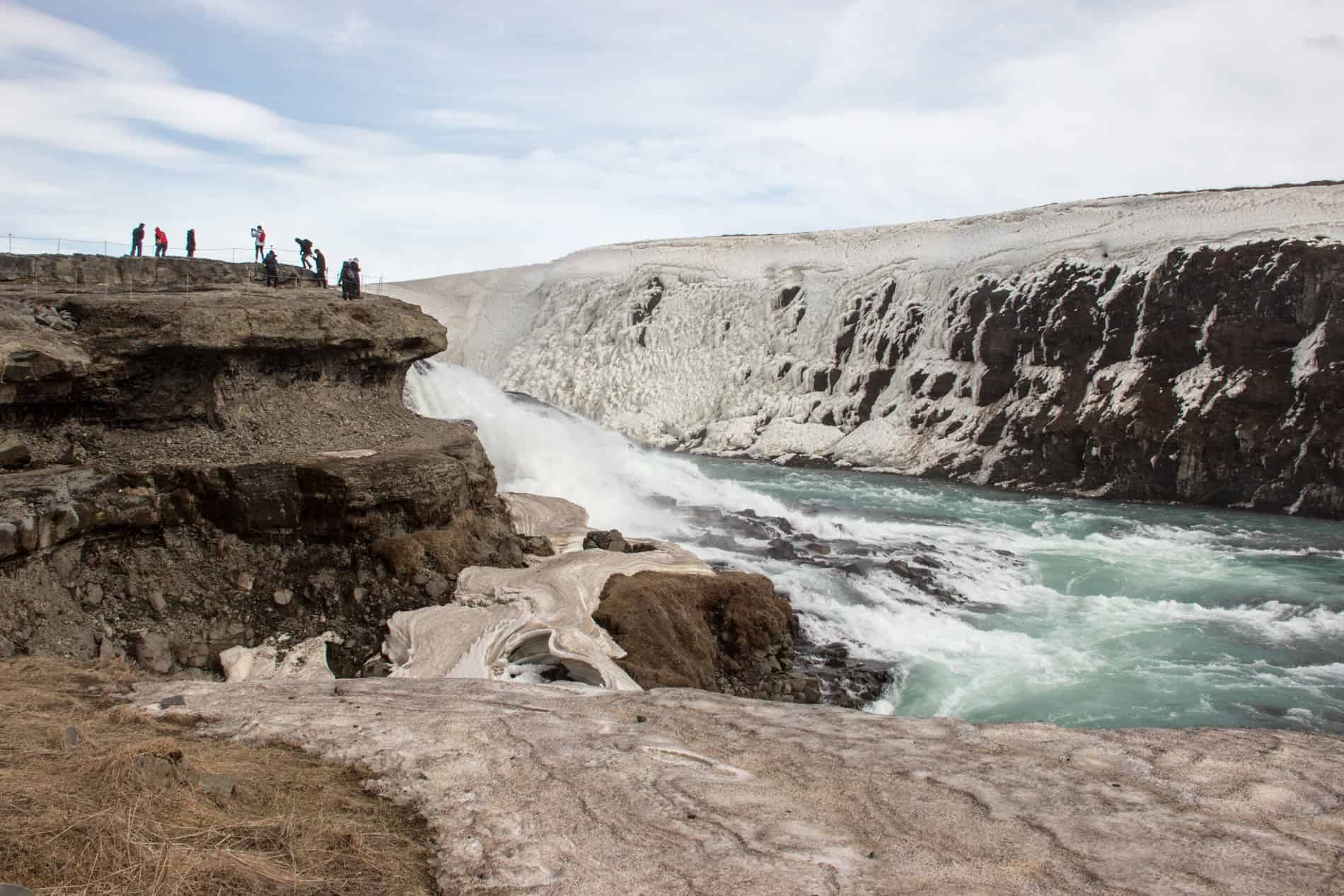 A cluster of visitors on a marked path on a rock ledge overlooking a powerful waterfall in Iceland.