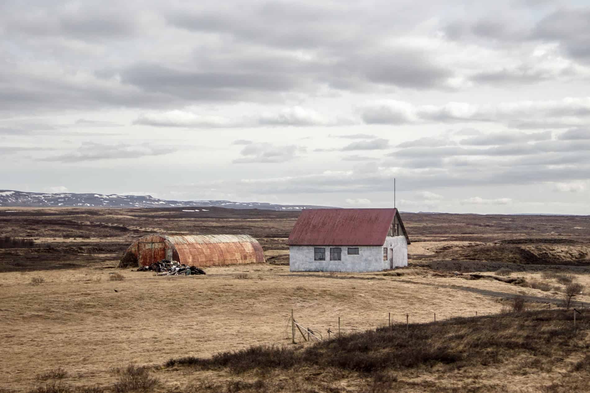 A farm house with a red roof and arched copper shed on flat, yellow land in Iceland. 