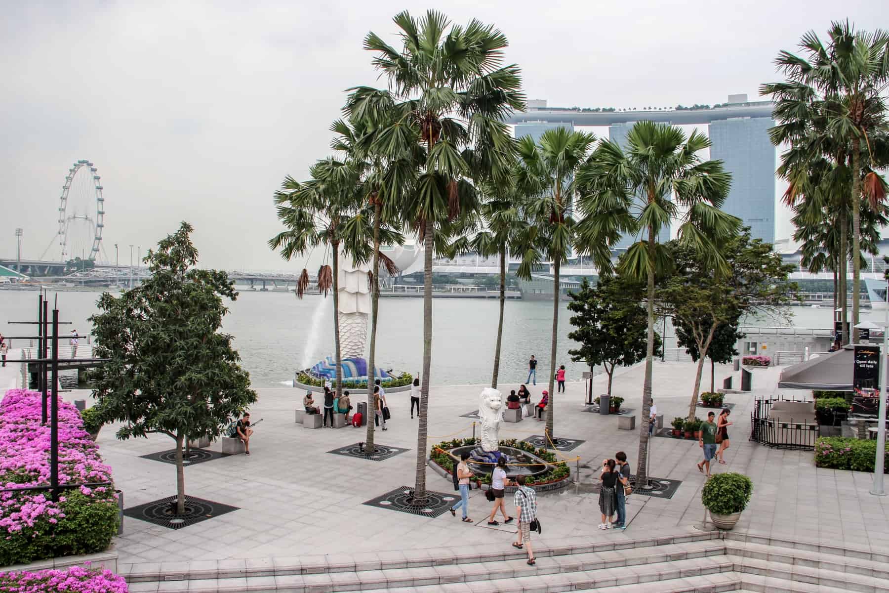 A waterside public park with green trees, pink flower bushes and the Merlion water fountain in Singapore looking out towards a ferris wheel and the boat shaped Marina Bays Sands hotel in the distance