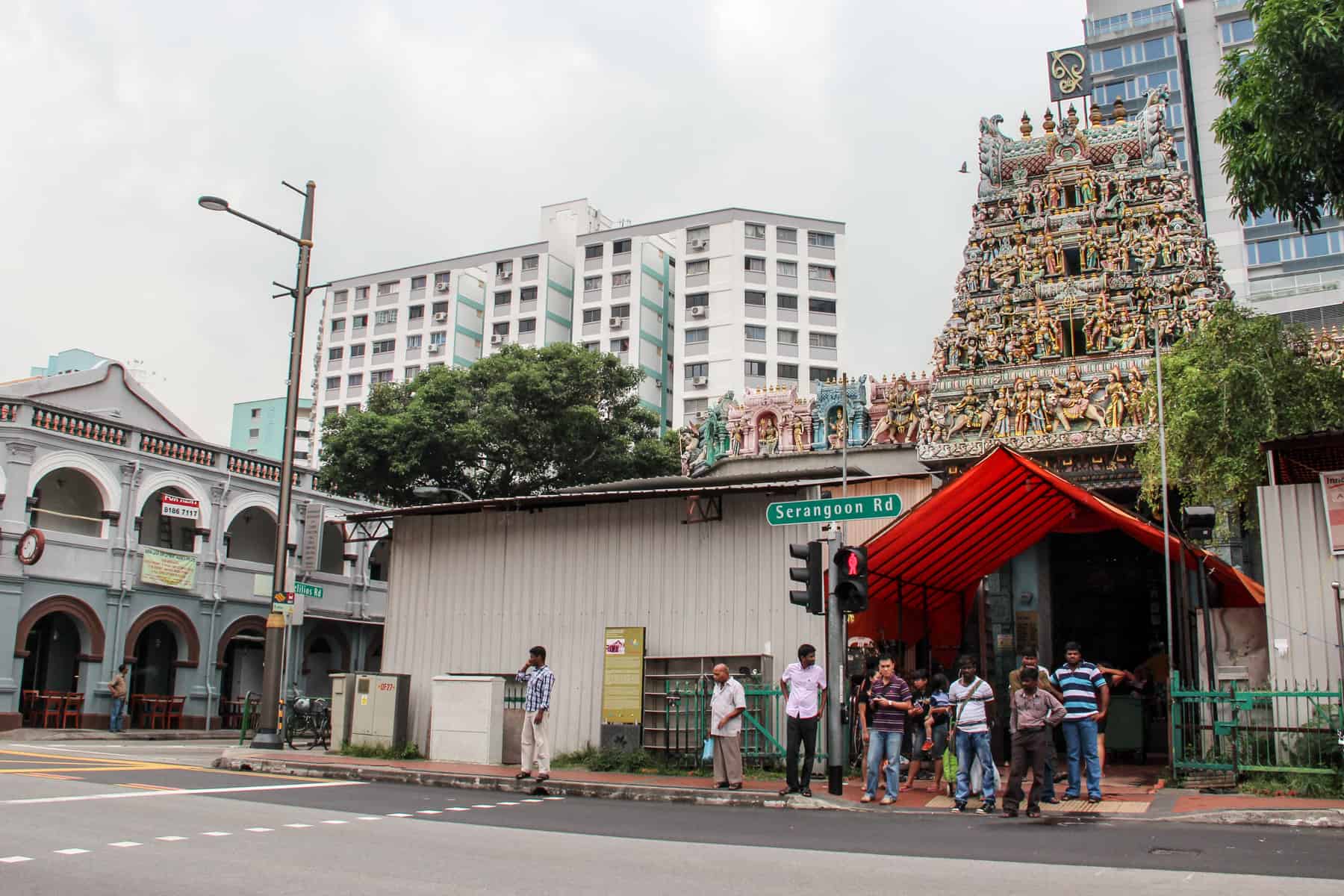 A small group of men at a road crossing right outside the entrance to the Sri Vadapathira Kaliamman temple in Singapore with a five-tier roof of pastel coloured deities and carvings