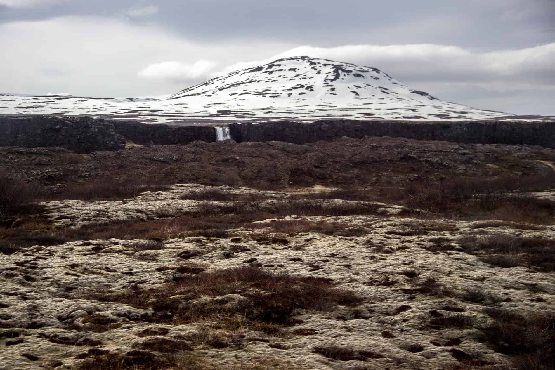 The uneven, rocky landscape of Pingvellir National Park, Iceland, backed by a small waterfall and a snow covered volcano. 