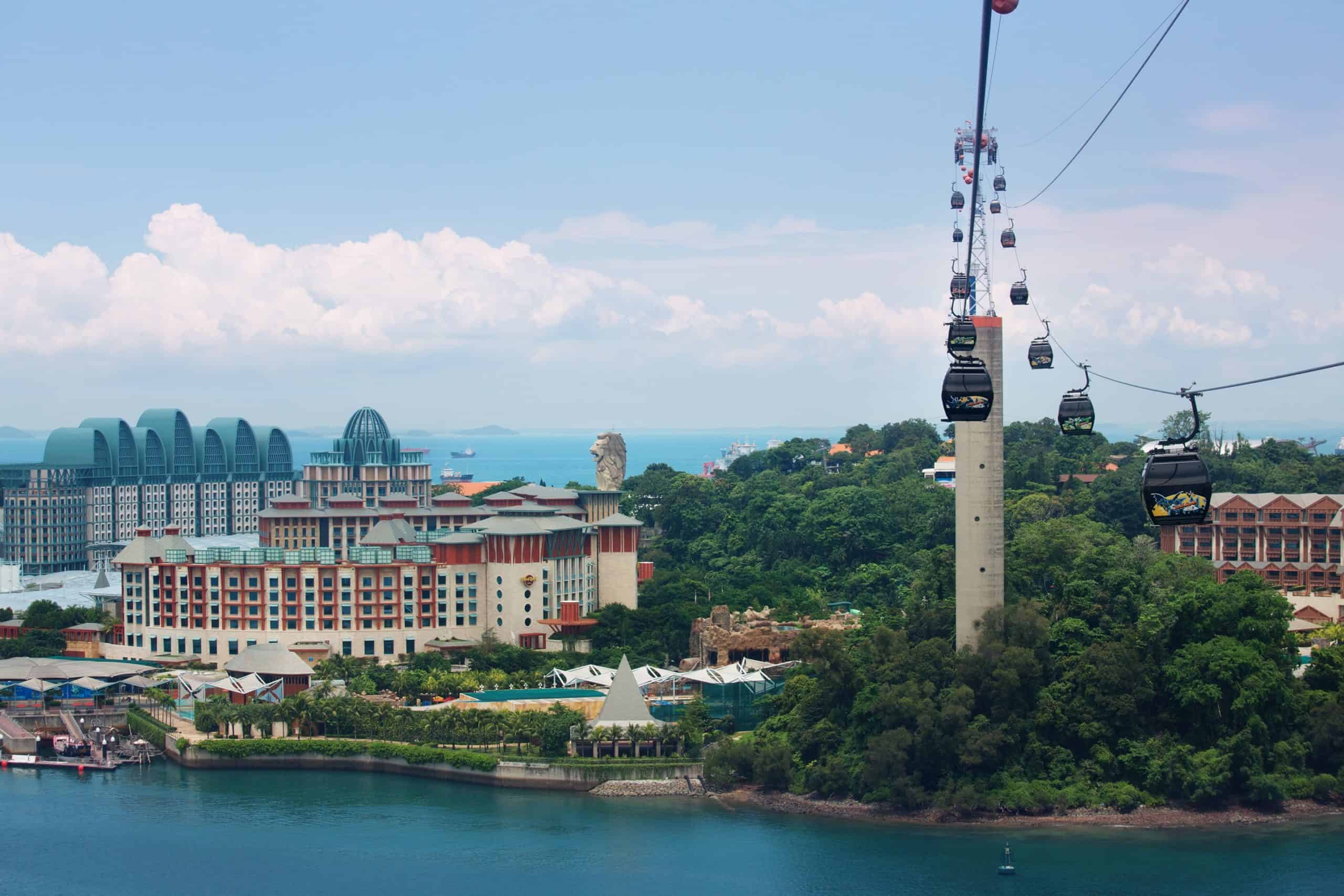 A long line of cables cars drift along the sky towards Sentosa Island in Singapore filled with big entertainment buildings and hotels