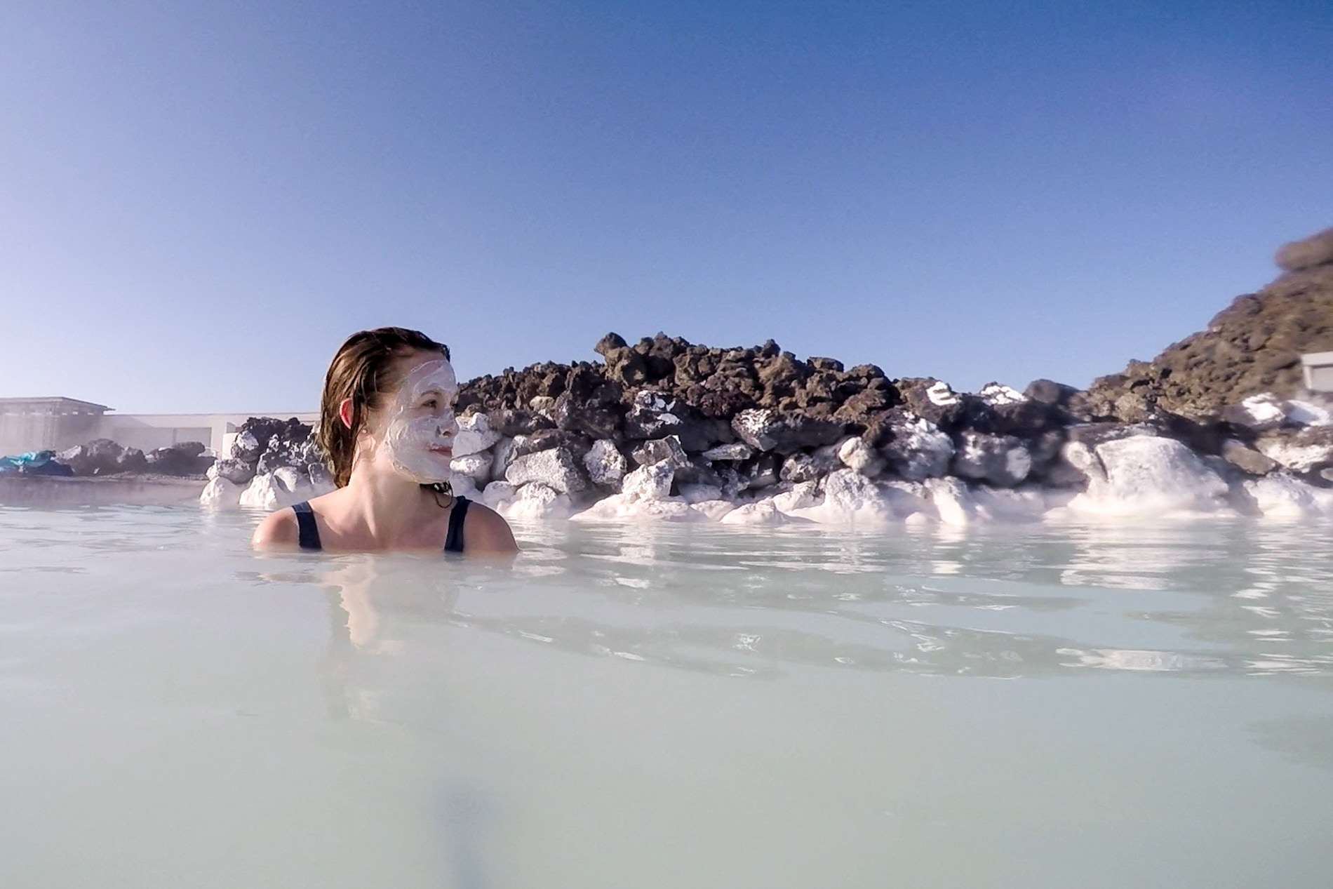 A woman, wearing a white clay face mask, looks ti the side as she submerges herself in the light waters of the Blue Lagoon in Iceland. Behind her are black rocks covered in a salt-like substance.