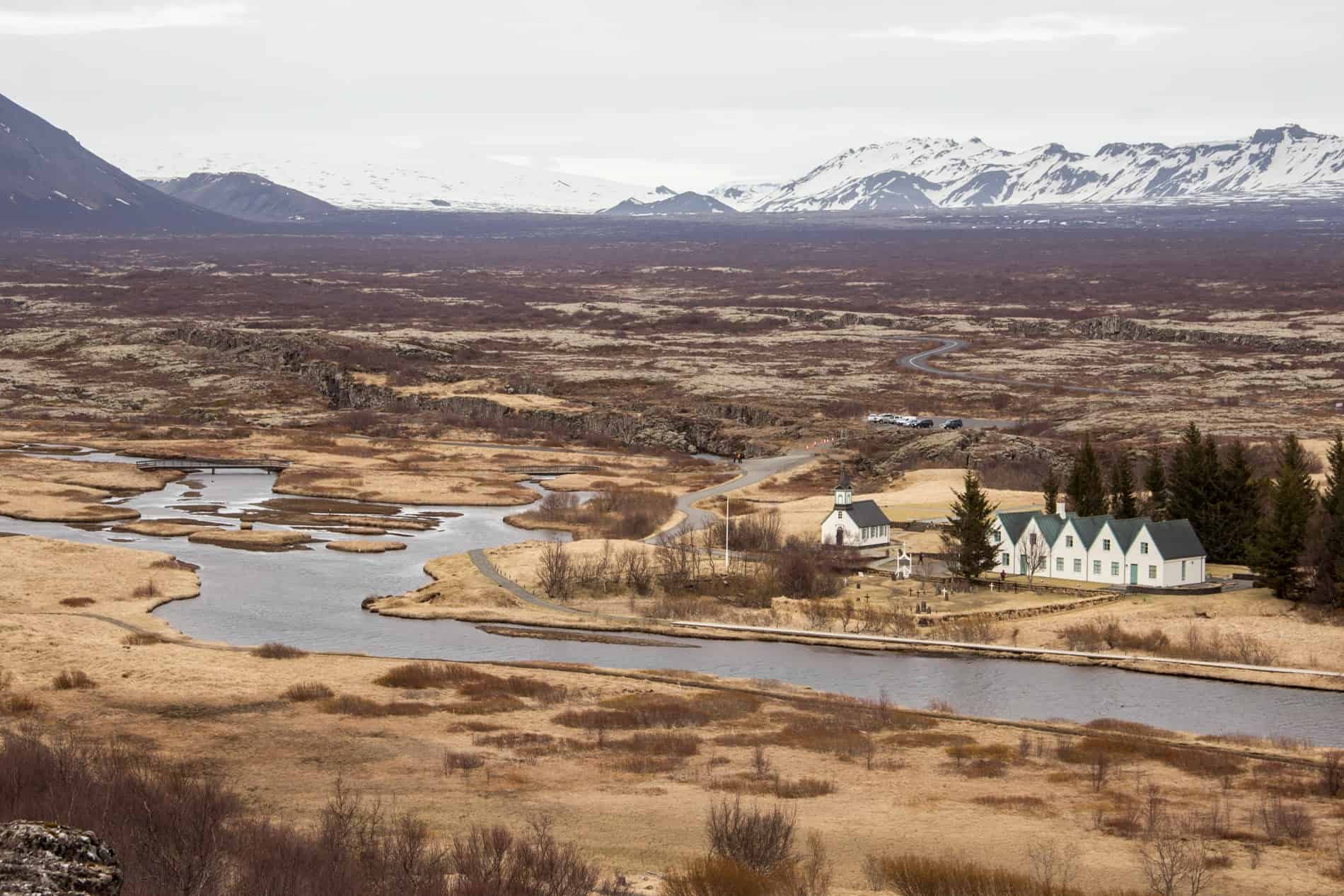 Six white triangular roof houses in a row, on dry sandy coloured landscape in front of a snowcapped mountain range. 