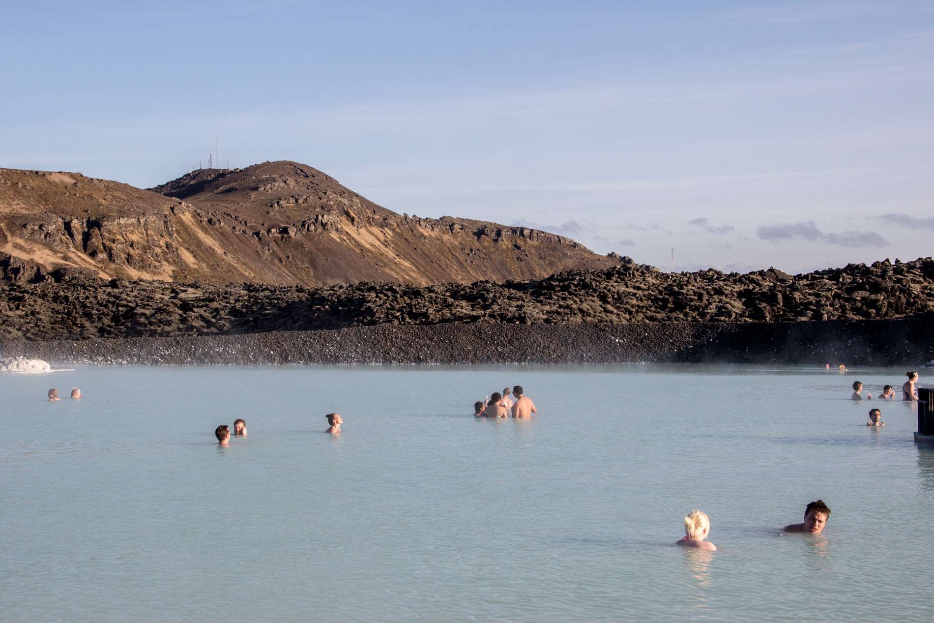People bathing in the bright blue waters of the Blue Lagoon in Iceland, backed by low rise, copper-coloured mountains. 