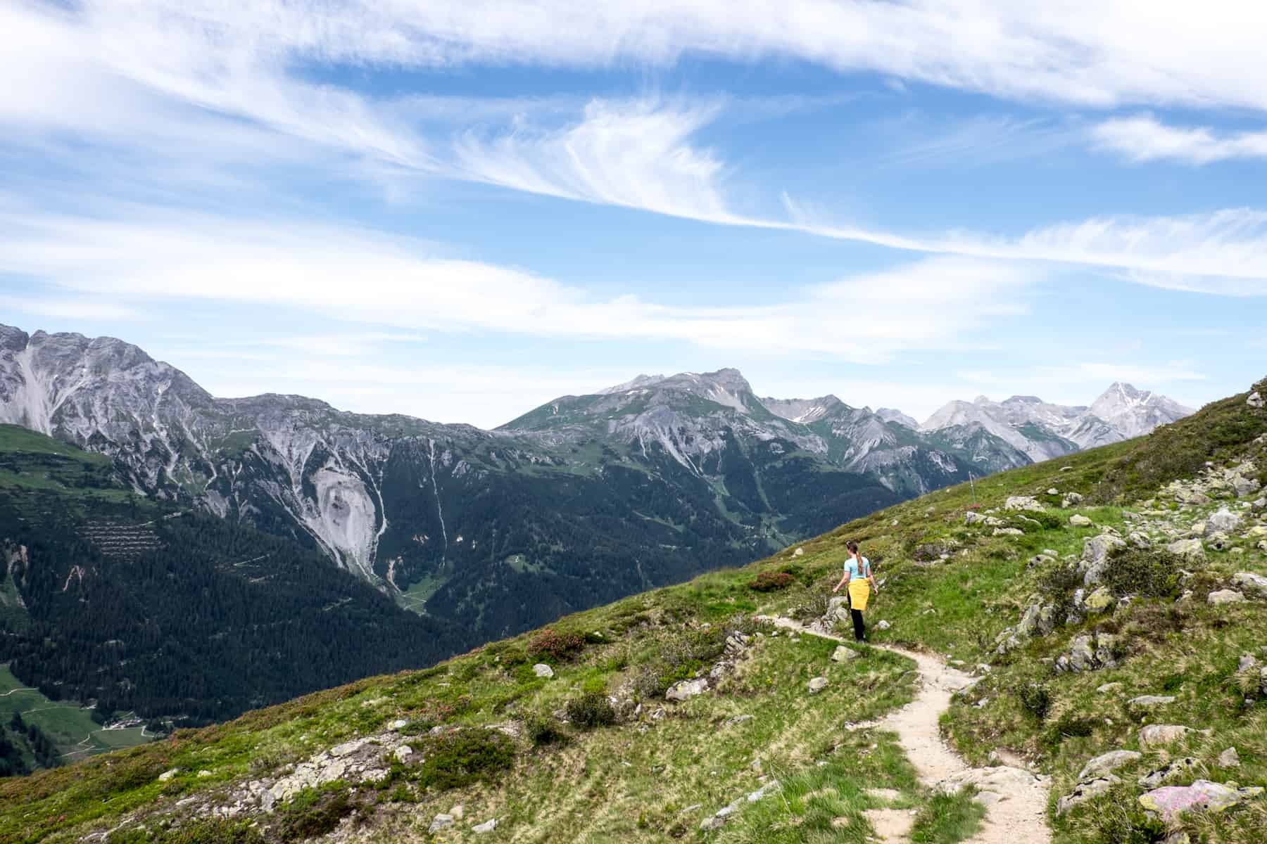 A woman hikes on a narrow hiking trail high on a green mountain in St. Anton. Snow-capped mountains can be seen in the background