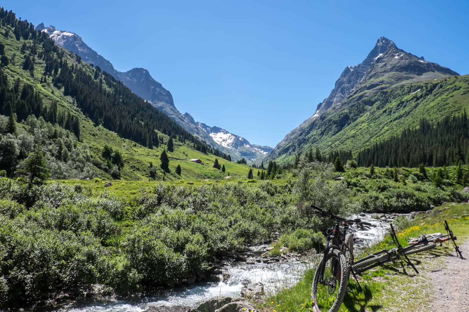 In St. Anton in Austria, two bikes rest next to a winding river that flows into a green valley, backed by mountains