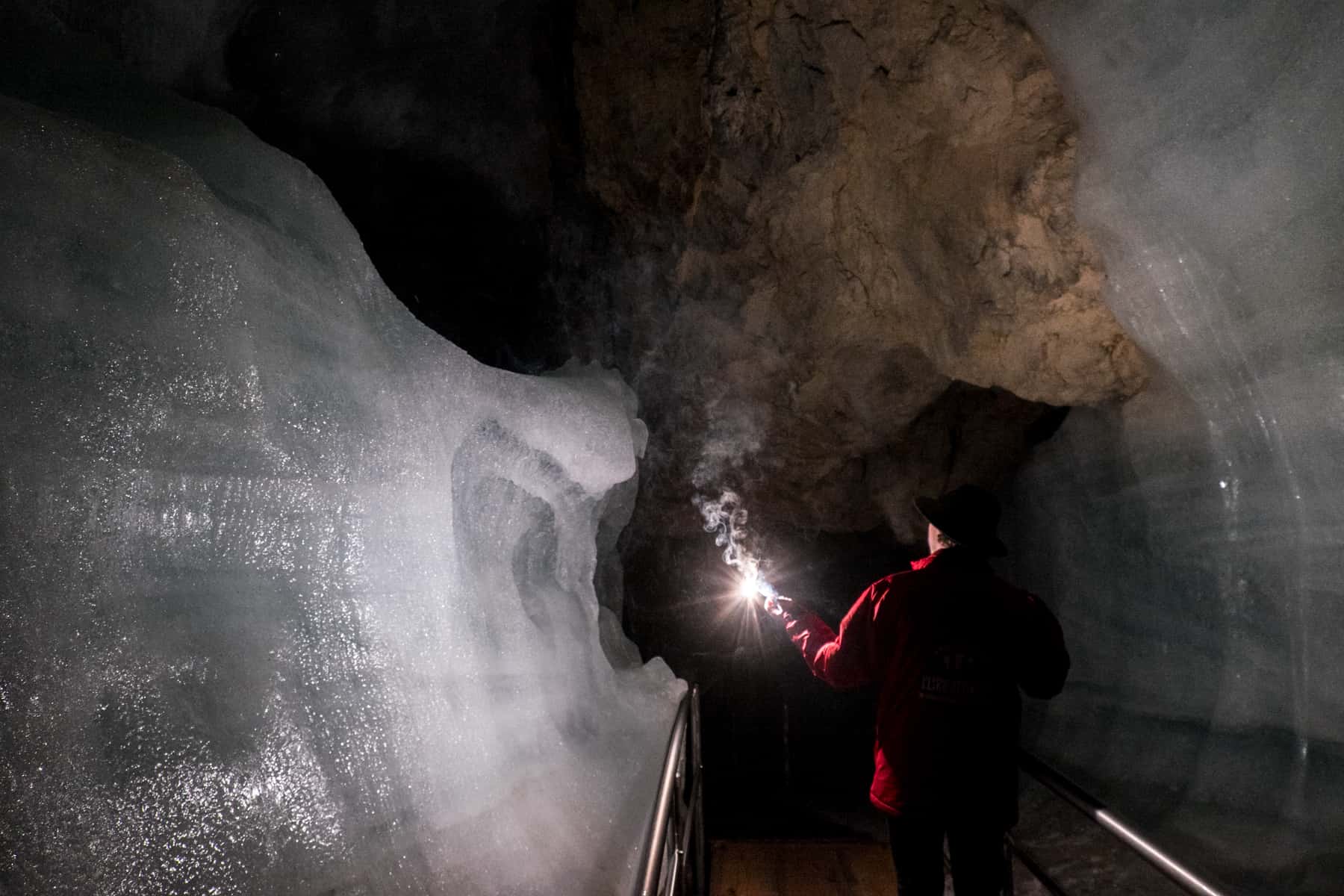 A man in a red jacket and black hat, holds a splint for light as he walks past a giant ice wall in Eisriesenwelt ice cave in Austria