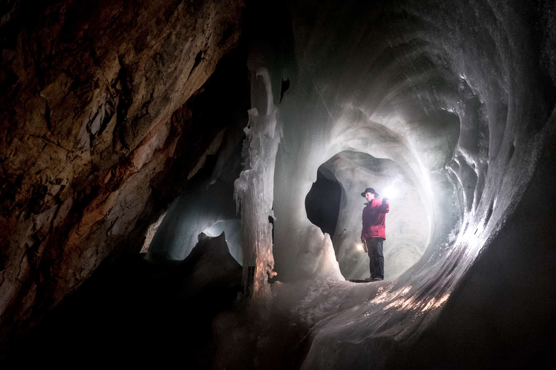 A man in a red jacket and black hat stands within a circle of white illuminated ice in the Eisriesenwelt largest ice cave in the world in Austria