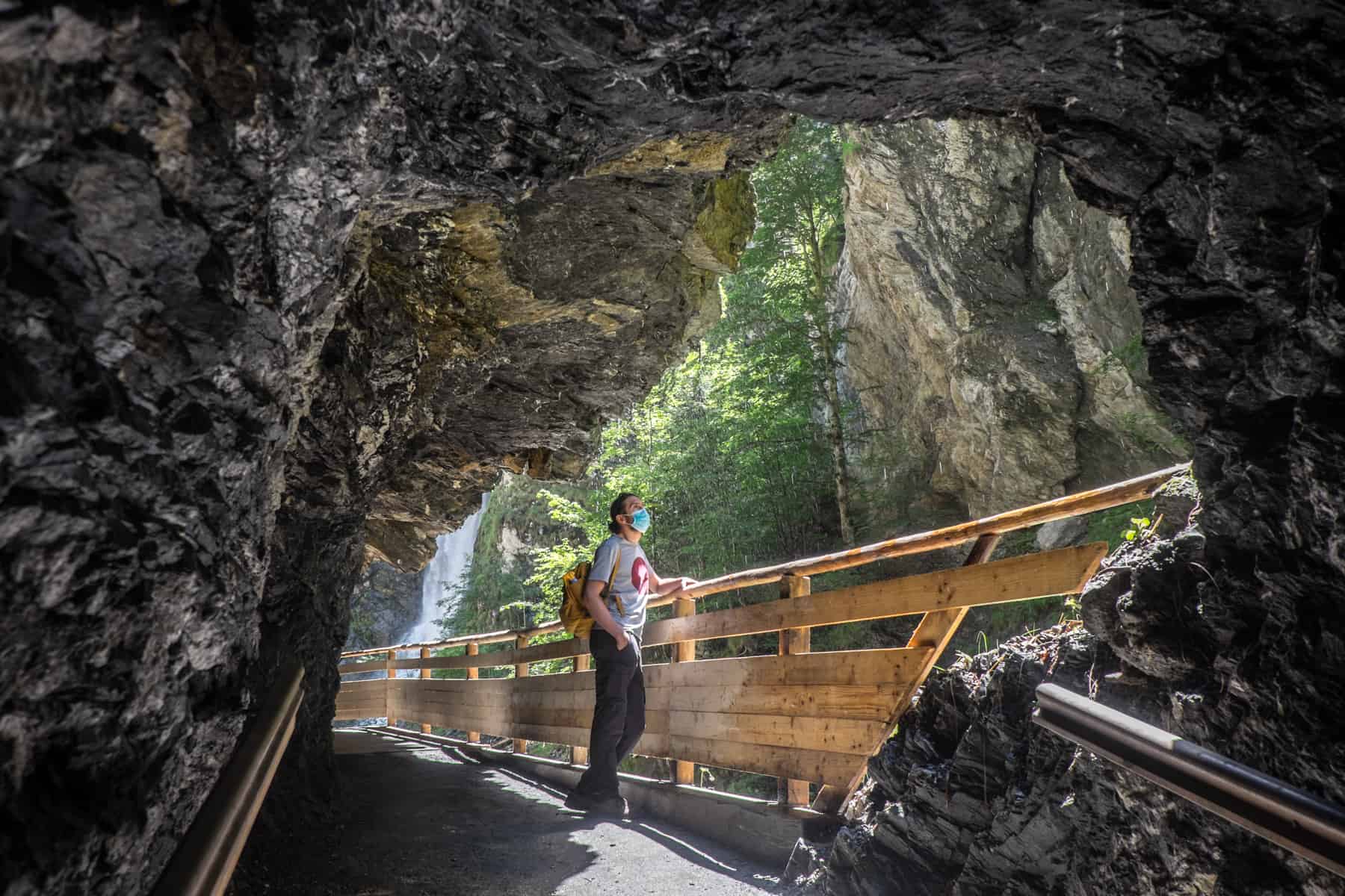 A man in a blue face mask, standing in a rocky archway, looks up out of the Liechtensteinklamm gorge while holding onto a wooden rail