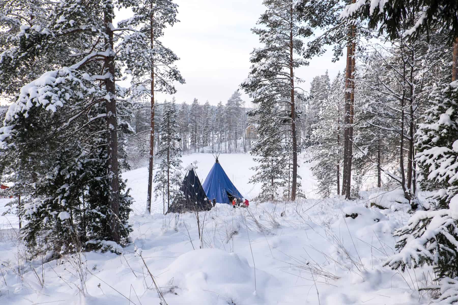 Two people dressed in red next to a black and blue teepee tent set within a forest covered in deep snow in Finland – where everyman has the right to roam. 